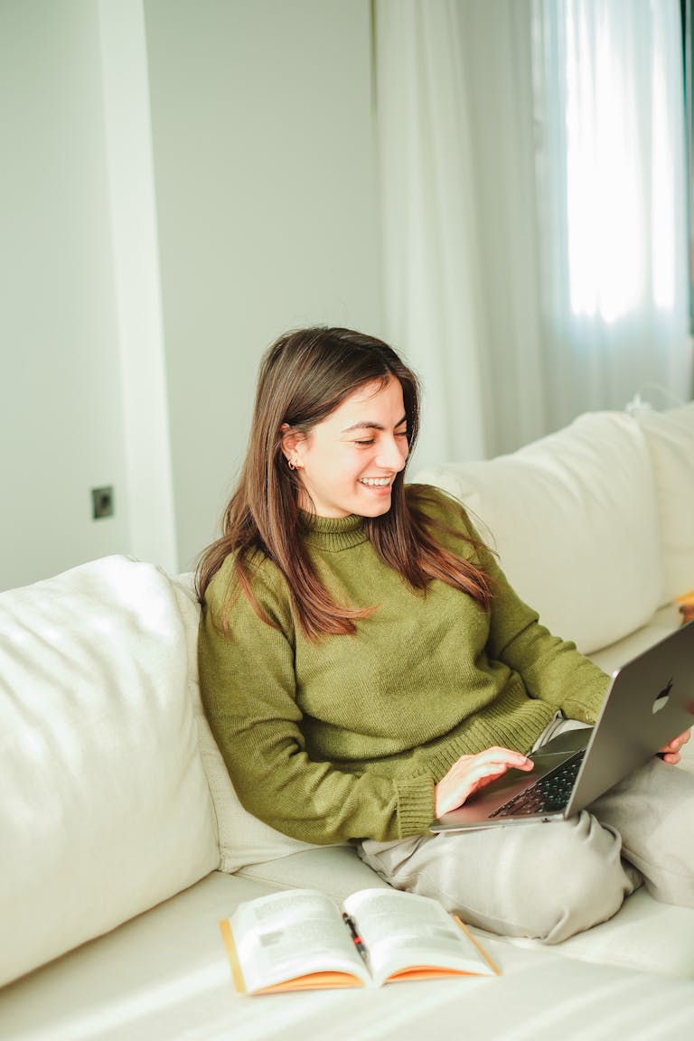 A young woman enjoys working on her laptop while relaxing on a sofa indoors, surrounded by books.